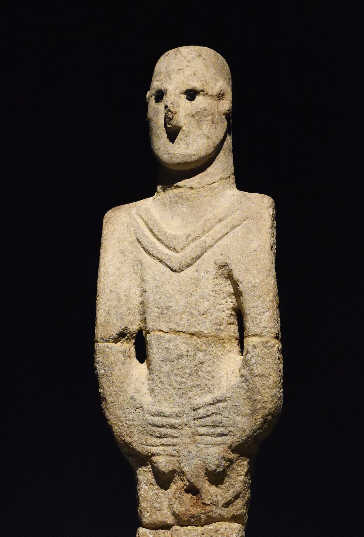 Limestone statue of a standing man with obsidian eyes, hands joined over the abdomen, displayed against a black background